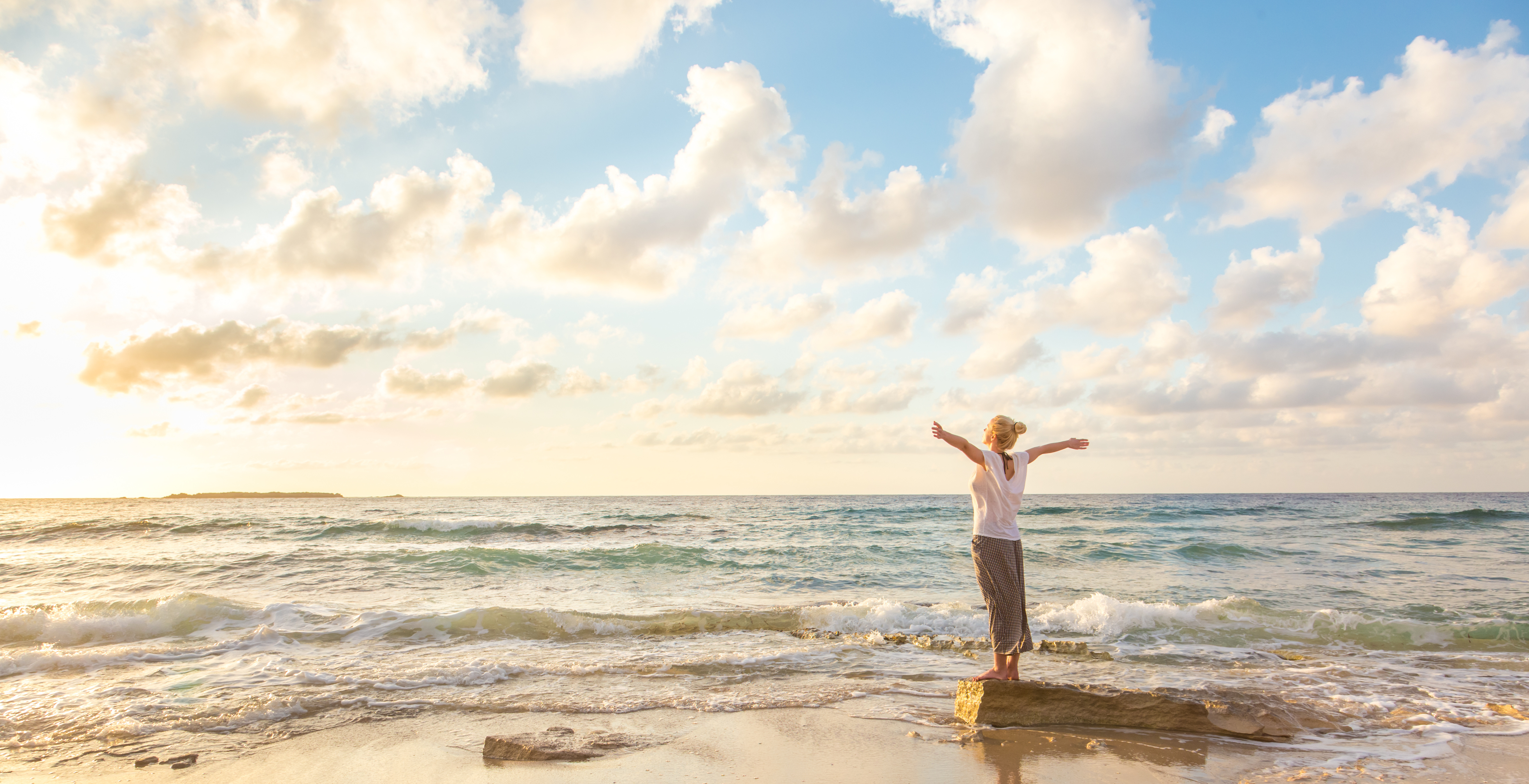 woman standing on rock in waves looking at ocean and sky with arms stretched out to sides